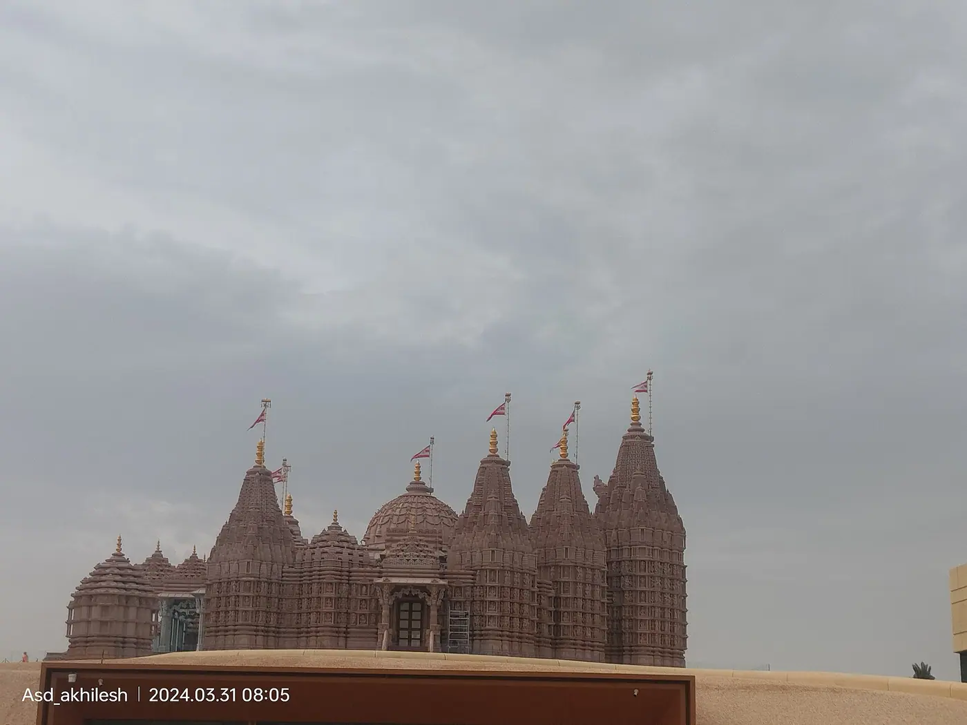 अबू धाबी में BAPS हिंदू मंदिर के गुलाबी बलुआ पत्थर के शिखर और गुंबद, केसरिया ध्वजों के साथ, बादलों से घिरे आकाश के नीचे रेत के टीले के ऊपर से दिखते हुए